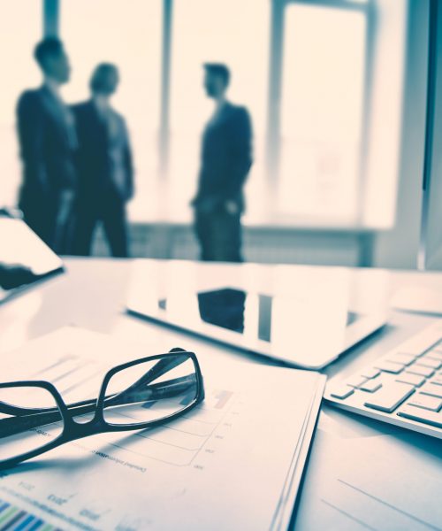 Modern devices, documents, eyeglasses on the foreground, unrecognized businessmen on the background